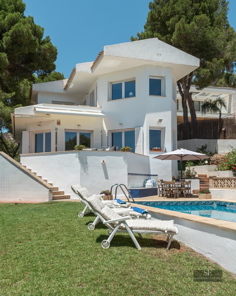 White two-story villa with a swimming pool, loungers, and green lawn surrounded by tall pine trees.