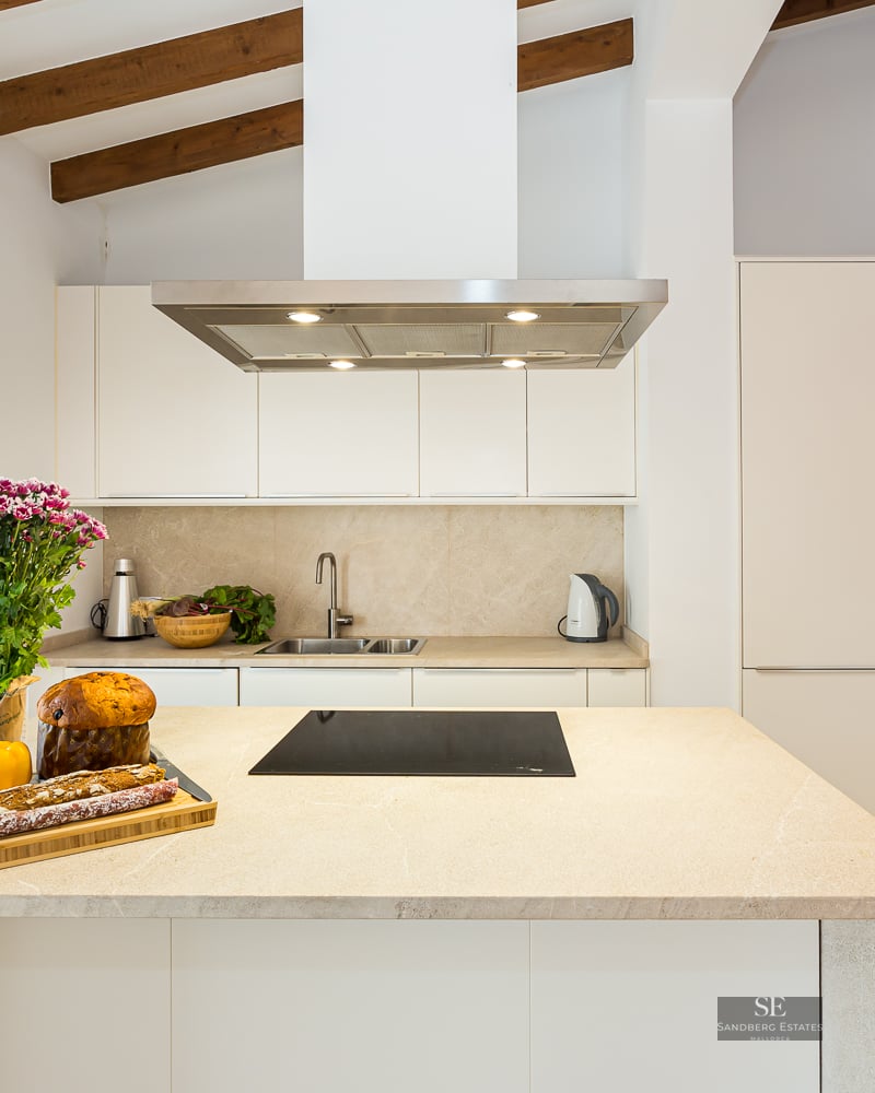 Bright kitchen featuring white cabinetry, a large stone island, rustic wooden ceiling beams, and integrated appliances.