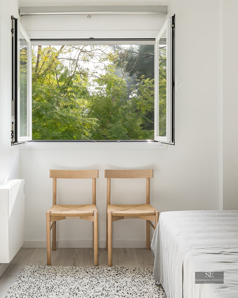 Bright minimalist bedroom with white walls, wooden chairs, grey bed, and an open window viewing lush green trees.