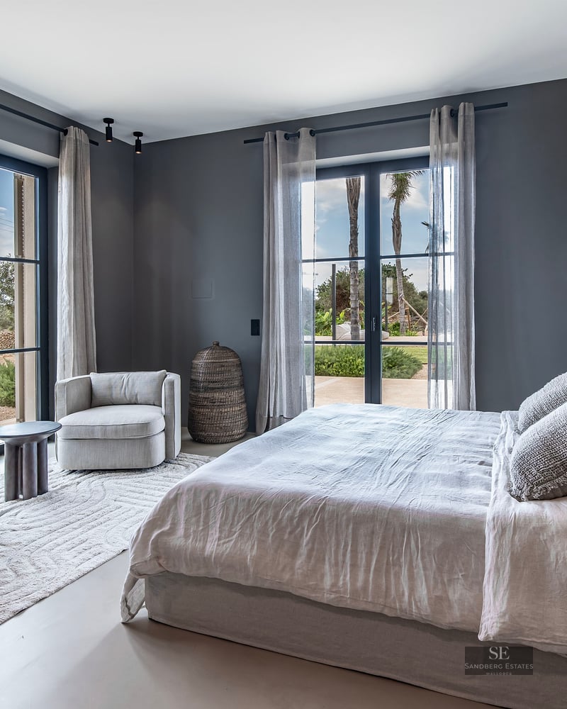 Elegant bedroom with grey walls, a large bed, armchairs, and floor-to-ceiling windows opening to a garden.