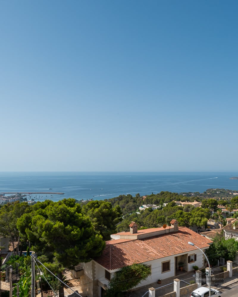 Panoramic hilltop view showing red-tiled roof houses, lush greenery, and the bright blue sea under a clear sky.