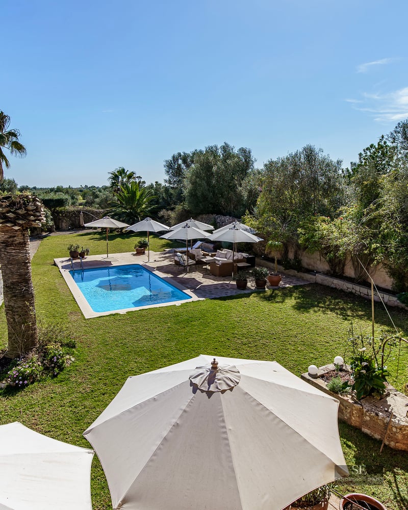 Elevated view of a blue swimming pool surrounded by green lawns, white umbrellas, and palm trees in a sunny garden.