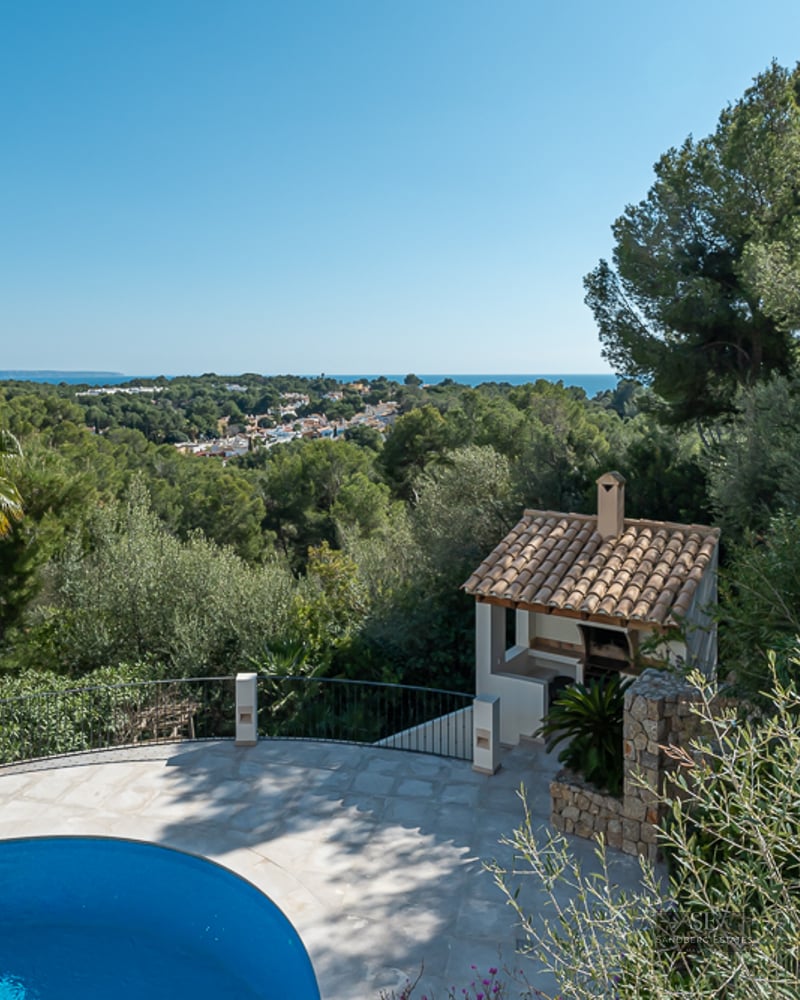 Piscine bleue incurvée sur une terrasse en pierre surplombant une vallée verdoyante et la mer au loin.