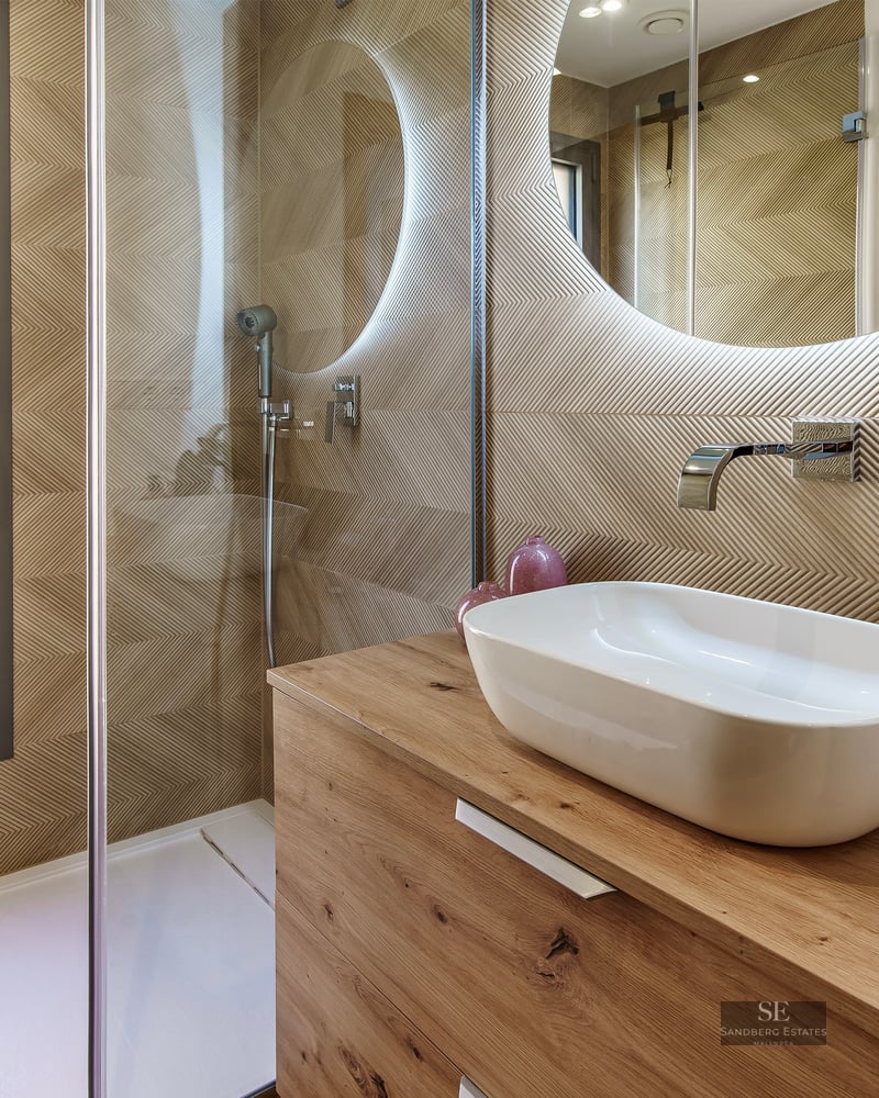 Modern bathroom featuring a wooden vanity, vessel sink, backlit round mirror, and a walk-in shower with glass screen.