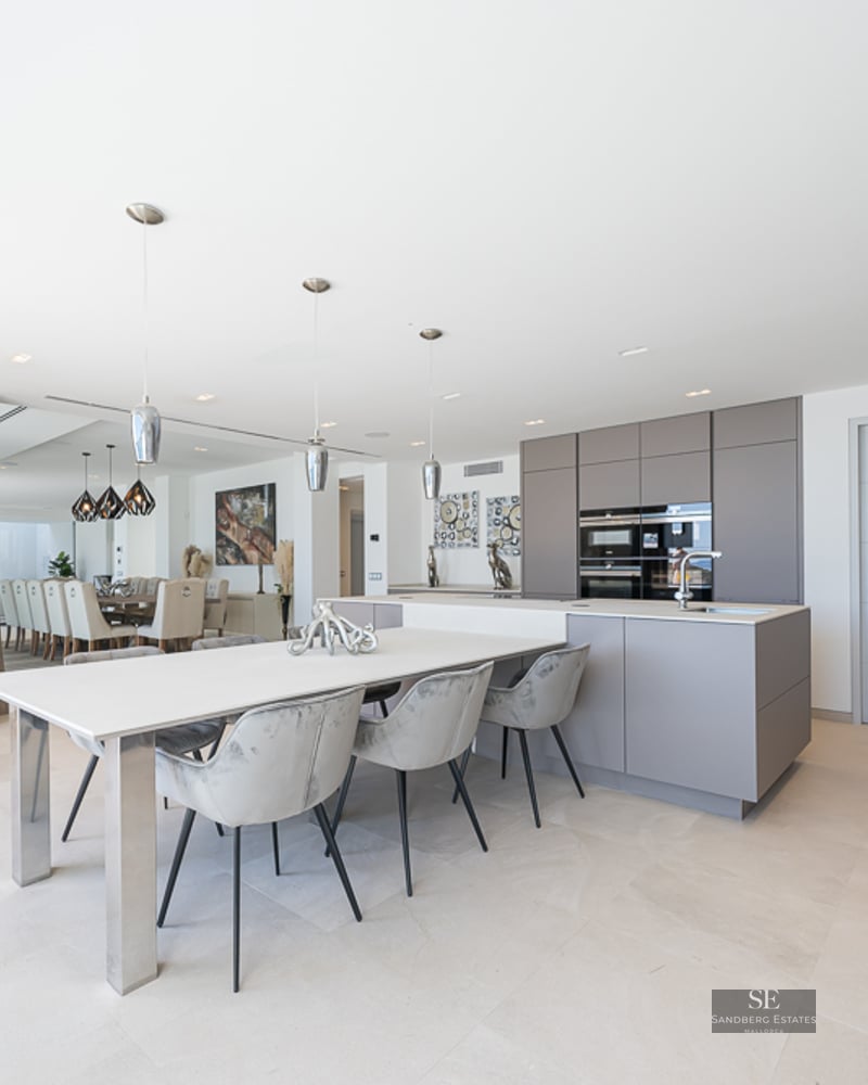 Modern kitchen with grey cabinetry, white island with attached dining table and grey velvet chairs.