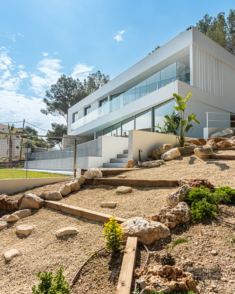 Side view of a modern two-story white villa with large glass windows and a tiered rock garden with gravel.