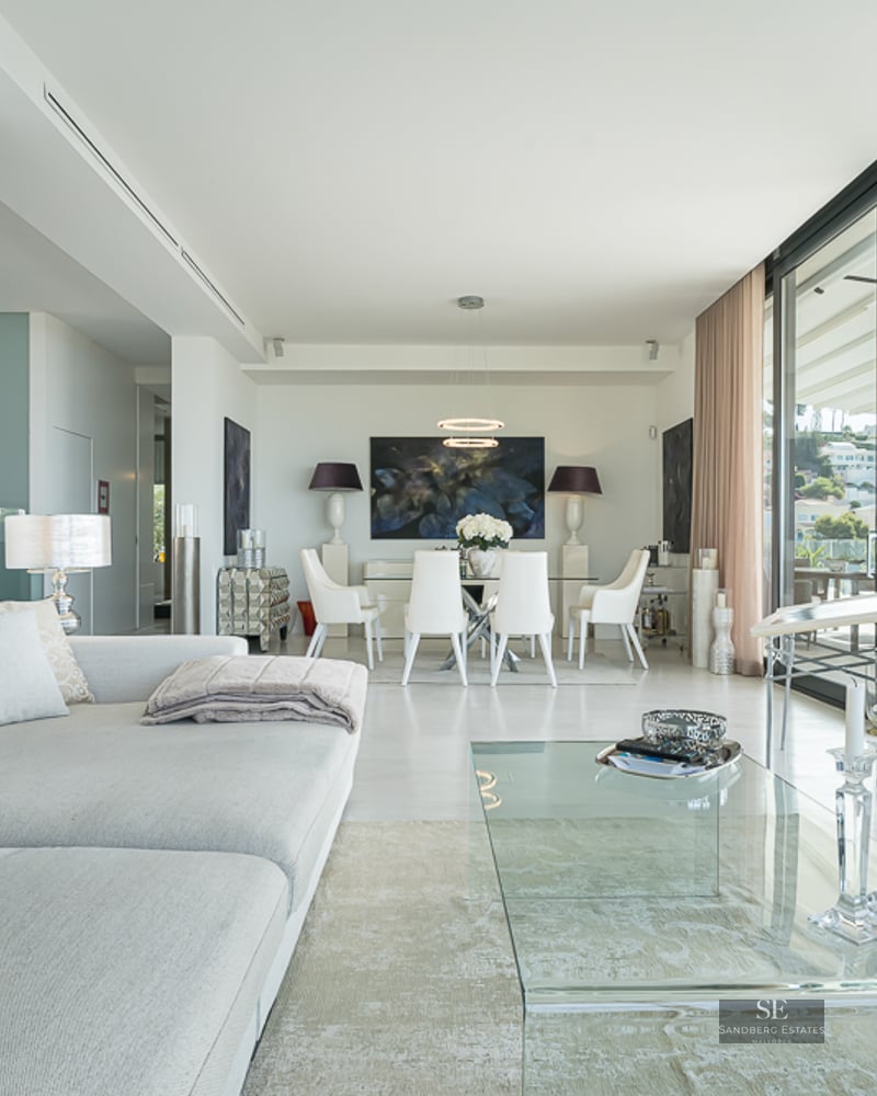 Bright, modern living room featuring a grey sectional sofa, glass coffee table, and floor-to-ceiling windows with views.