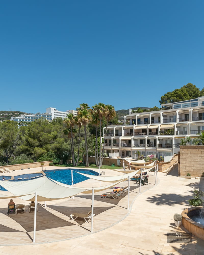 A large blue swimming pool surrounded by a stone terrace with white shade sails and palm trees under a clear blue sky.