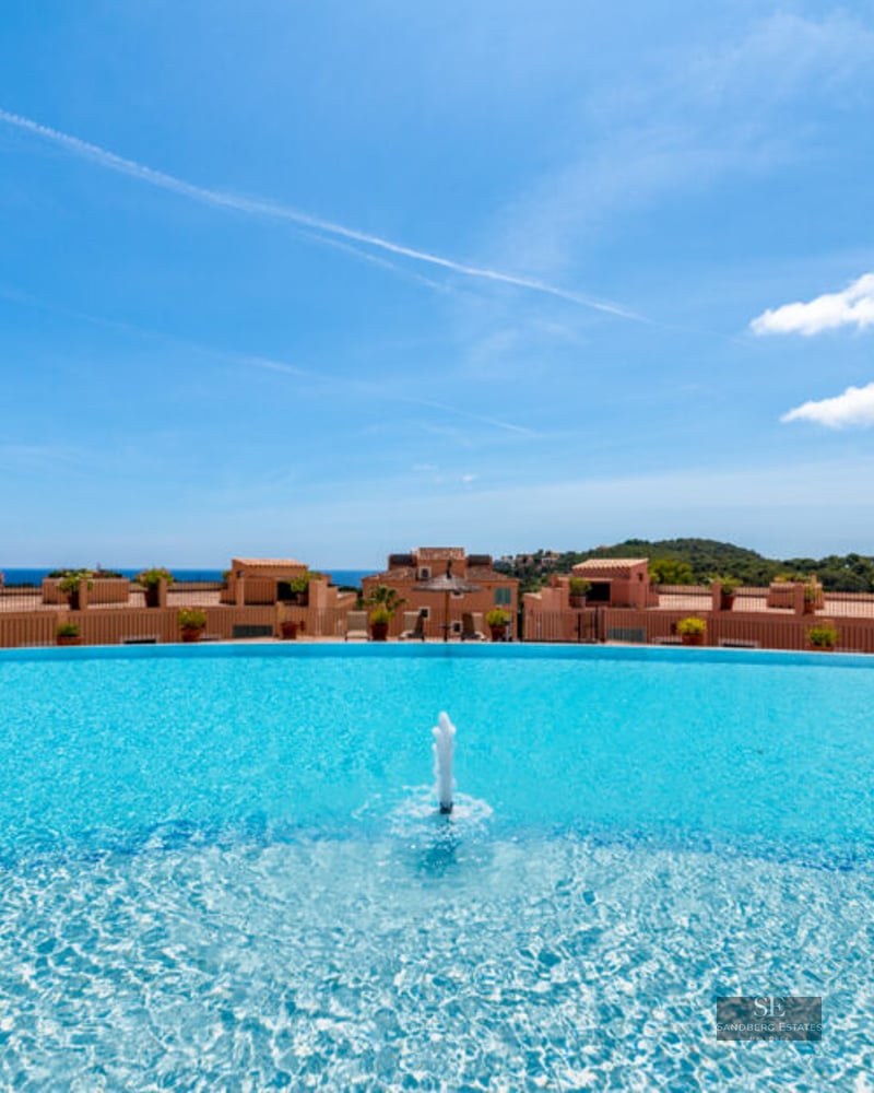 Grande piscine incurvée avec fontaine centrale et vue panoramique sur les bâtiments méditerranéens et la mer.