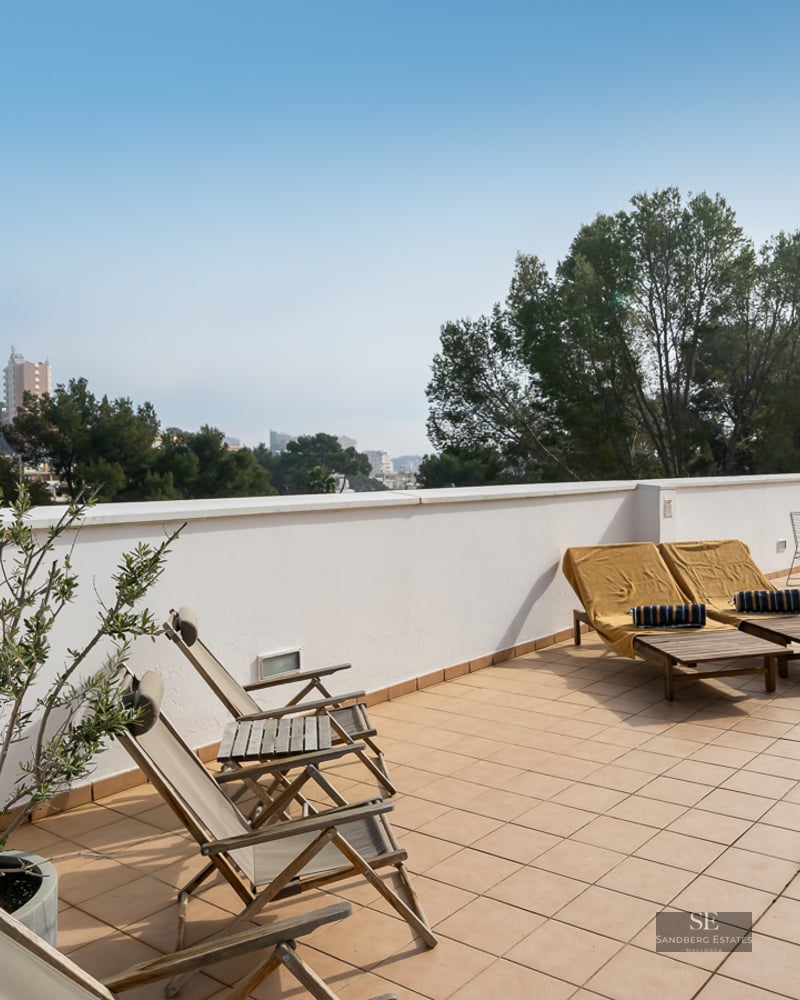 Large tiled terrace with wooden lounge chairs, potted plants, and a view of trees and city buildings.
