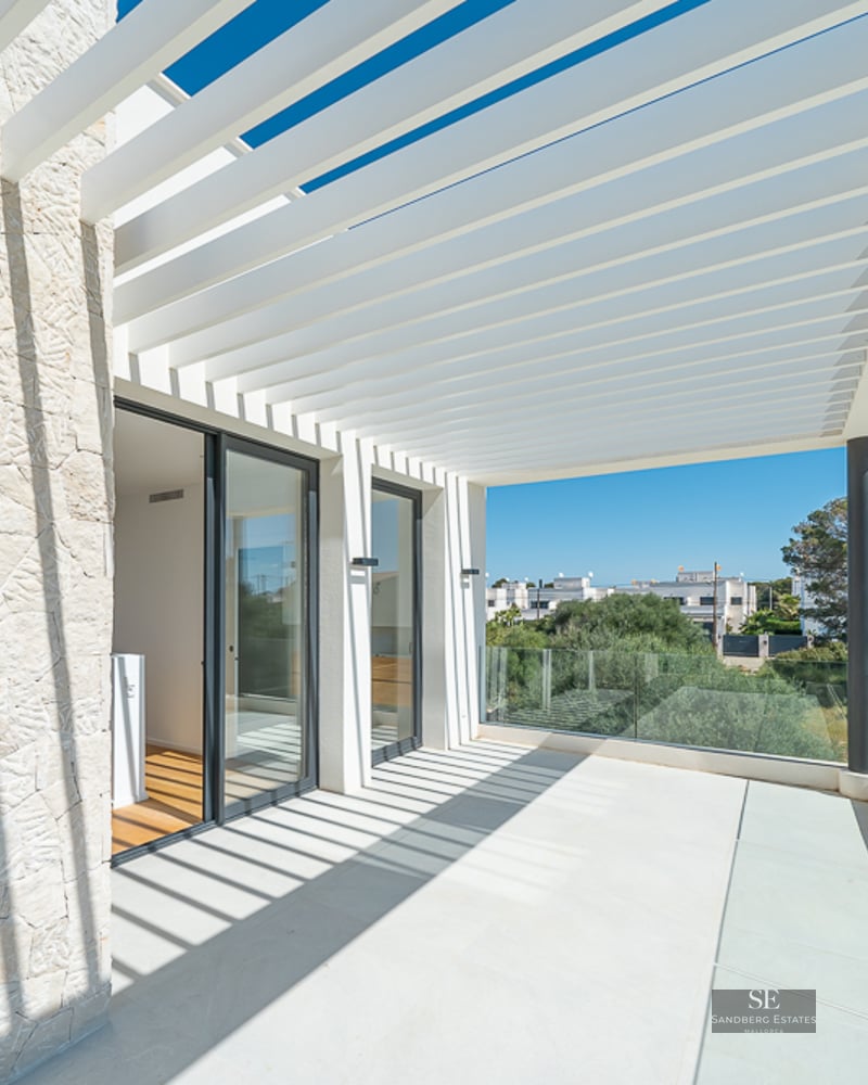 Bright outdoor terrace with a white slatted pergola casting shadows on a textured stone wall and glass railing.