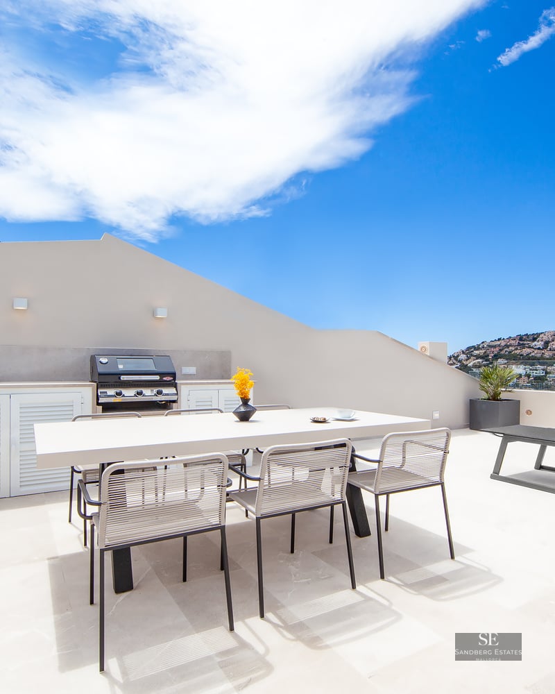 Modern rooftop terrace featuring an outdoor kitchen, dining table, and sun loungers with panoramic coastal views.