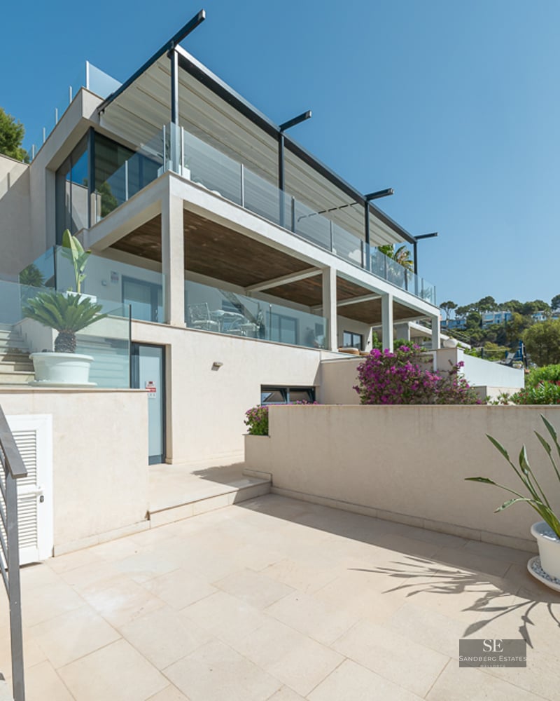 Multi-story modern villa with beige stone stairs, glass railings, and bougainvillea plants under a clear blue sky.