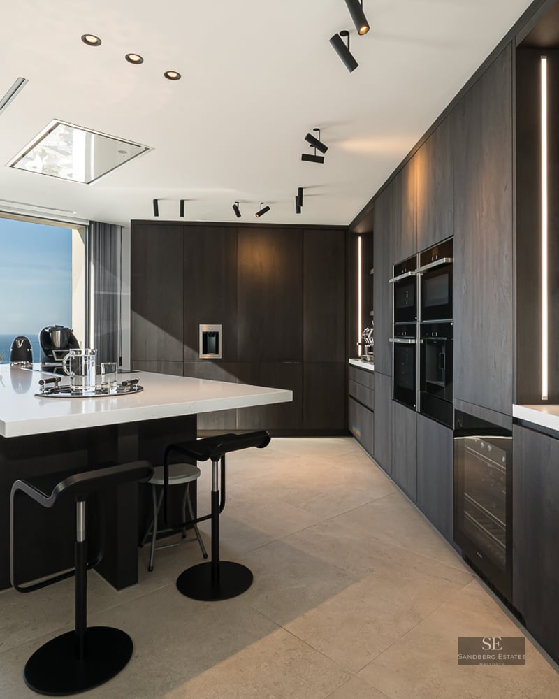 Contemporary dark wood kitchen with a large white island and panoramic ocean views through floor-to-ceiling windows.