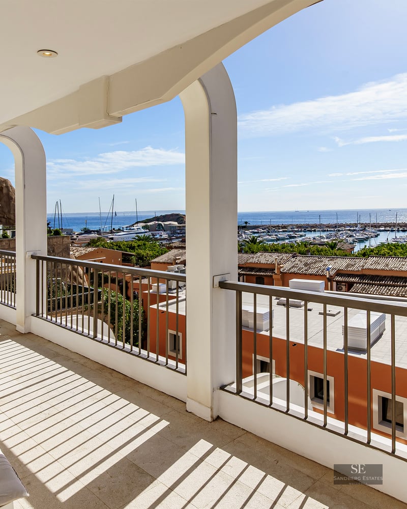 White arched balcony with outdoor furniture overlooking a harbor with boats and blue sea under a clear sky.