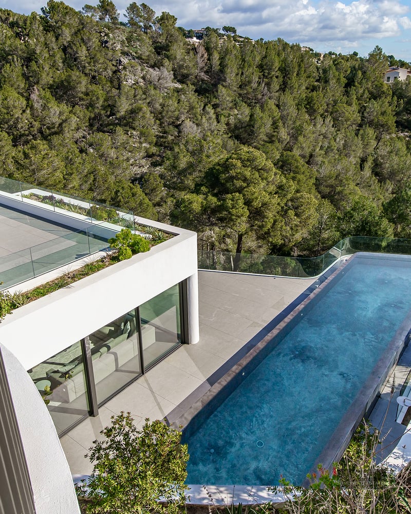 A high-angle view of a modern villa's infinity pool overlooking a lush green forest landscape under a clear sky.