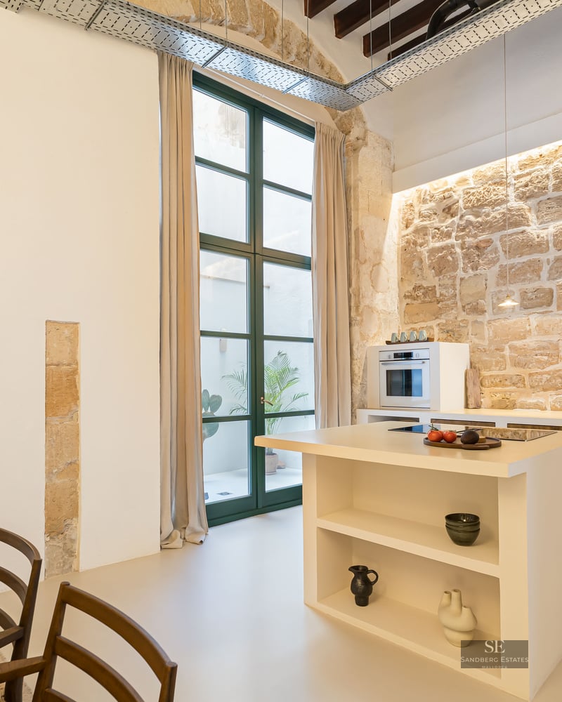 Contemporary kitchen featuring a rustic stone wall, white kitchen island, high ceilings with wooden beams and a tall window.