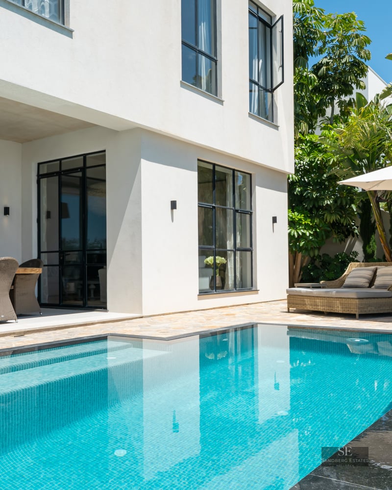 Luxury swimming pool next to a white villa with an outdoor dining area, daybed, and tropical garden.
