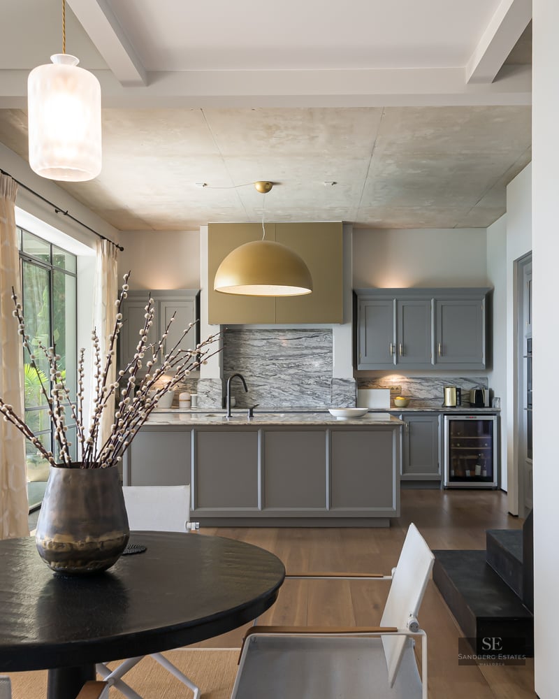 Modern kitchen with grey island, marble backsplash, gold lamp, and dining table with chairs by a large window.