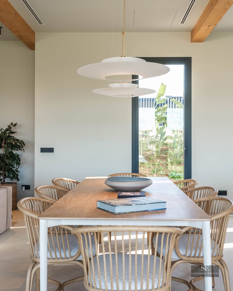 Modern dining room featuring a wooden table, rattan chairs, a designer pendant light, and a window with garden views.