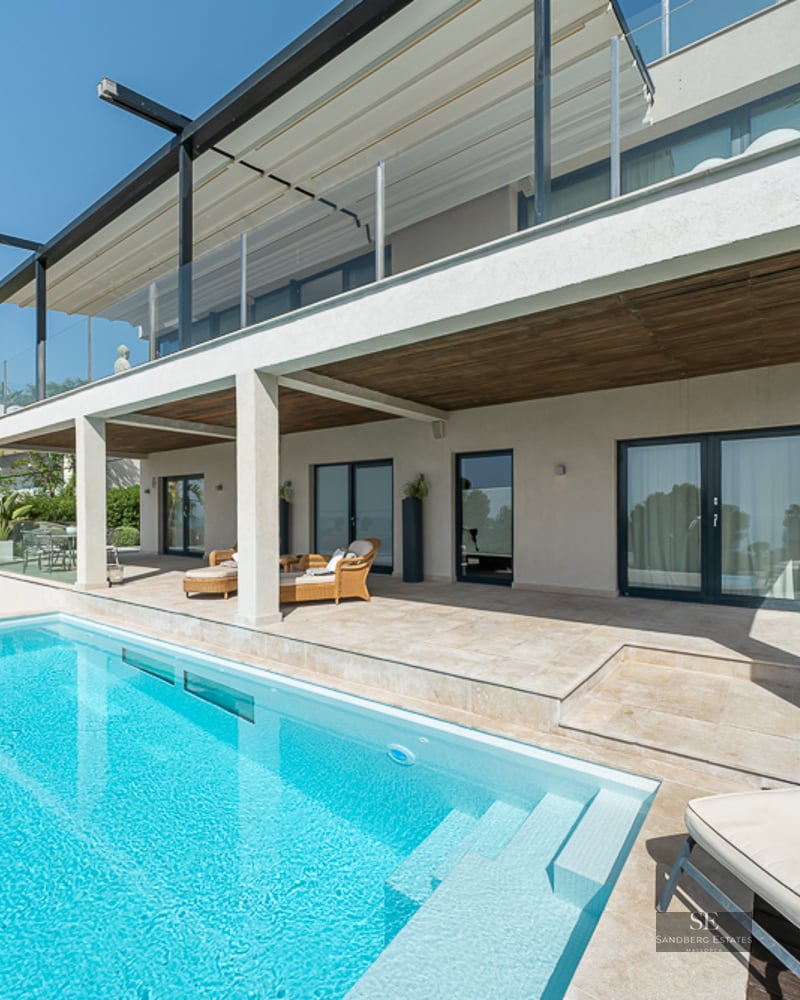 Crystal clear blue swimming pool adjacent to a stone terrace with wicker furniture and a modern white villa.