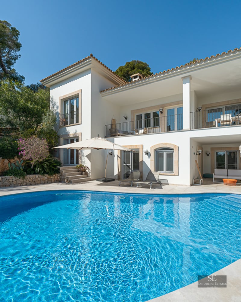 Grande piscine bleue devant une villa méditerranéenne blanche à deux étages avec terrasses et arbres.
