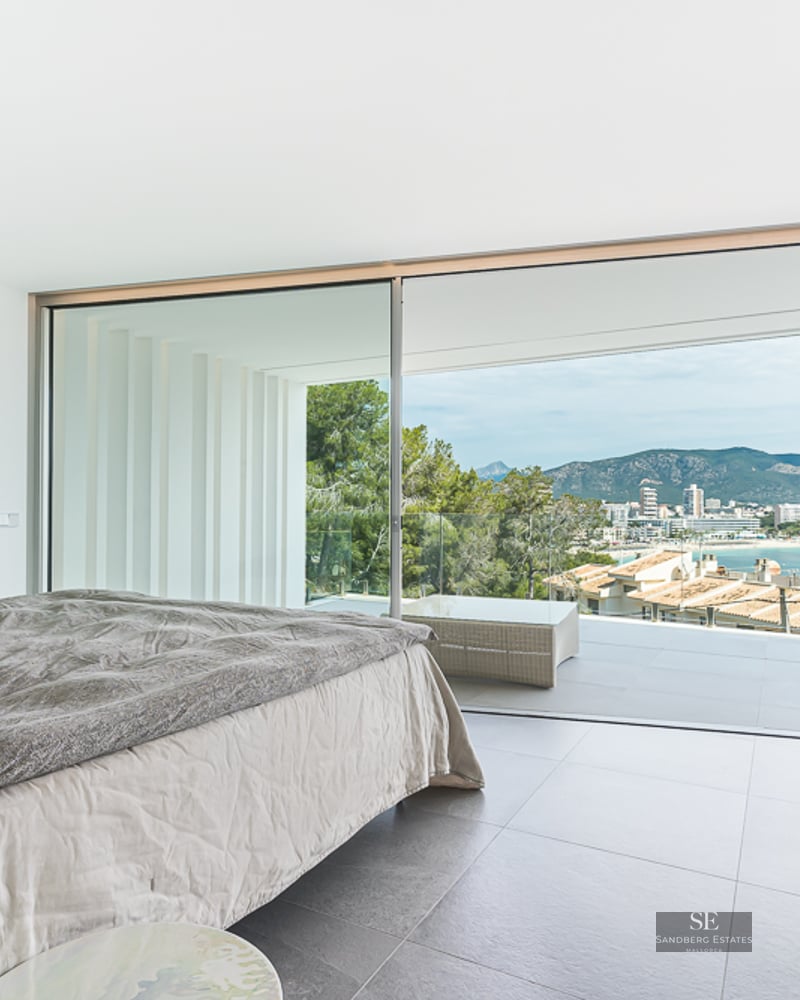Bright modern bedroom with a large bed and floor-to-ceiling glass doors opening to a terrace with coastal views.