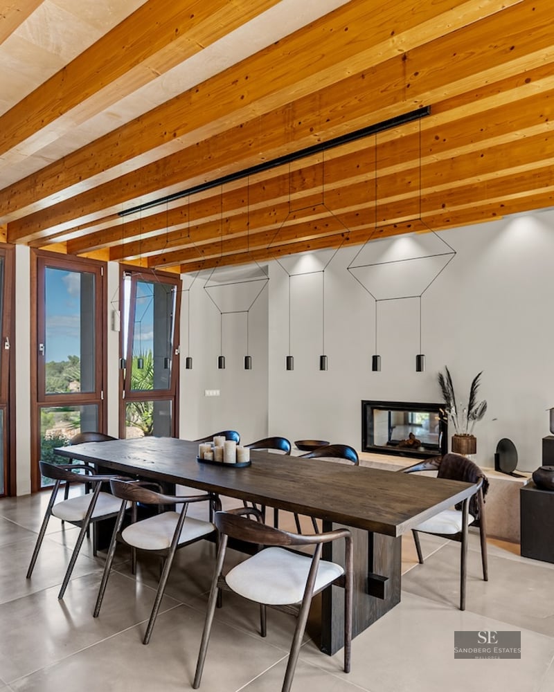 Large wooden dining table under a beamed ceiling with floor-to-ceiling windows showing nature views.