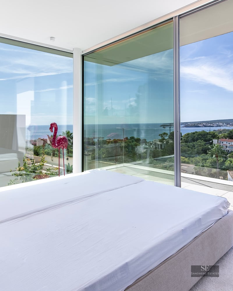 Modern bedroom with floor-to-ceiling corner windows overlooking the Mediterranean sea and coastal landscape.