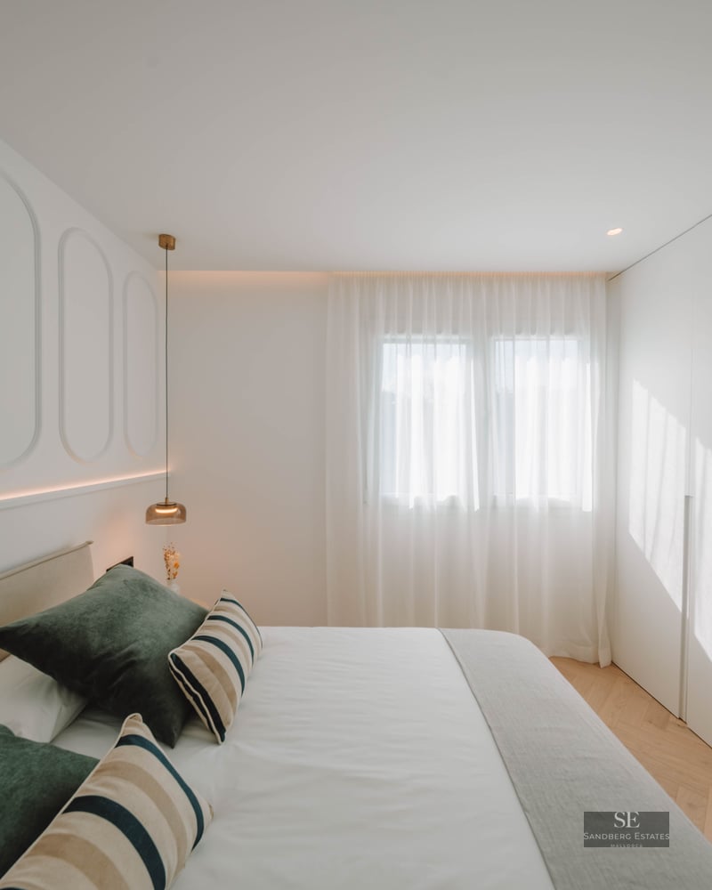 Bright bedroom with white paneled walls, a double bed with green pillows, herringbone wood floors, and built-in closets.