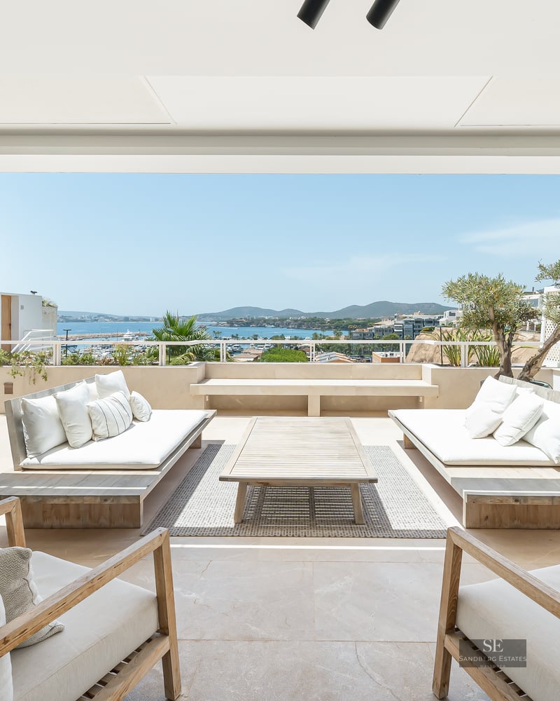 Spacious white terrace with wooden sofas and armchairs overlooking a blue sea and coastline under a bright sky.