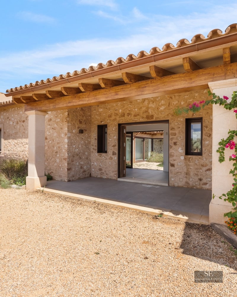 Entrance of a stone villa with a wooden porch, gravel path, and vibrant flowers under a blue sky.