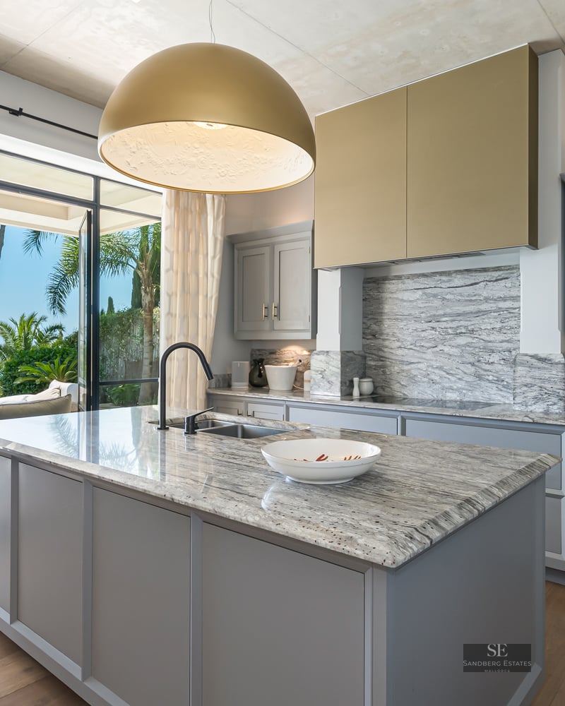 Modern kitchen with a marble island, grey cabinets, and a large open window overlooking the Mediterranean sea.