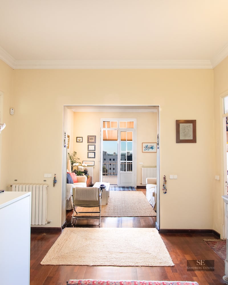 Hallway with wood floors leading to a bright living room with sofas and a door to a sunny outdoor balcony.