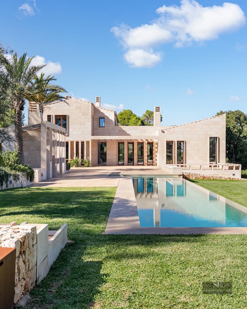Modern sandstone villa with a long rectangular lap pool, lush green lawn, and ancient olive trees under a blue sky.
