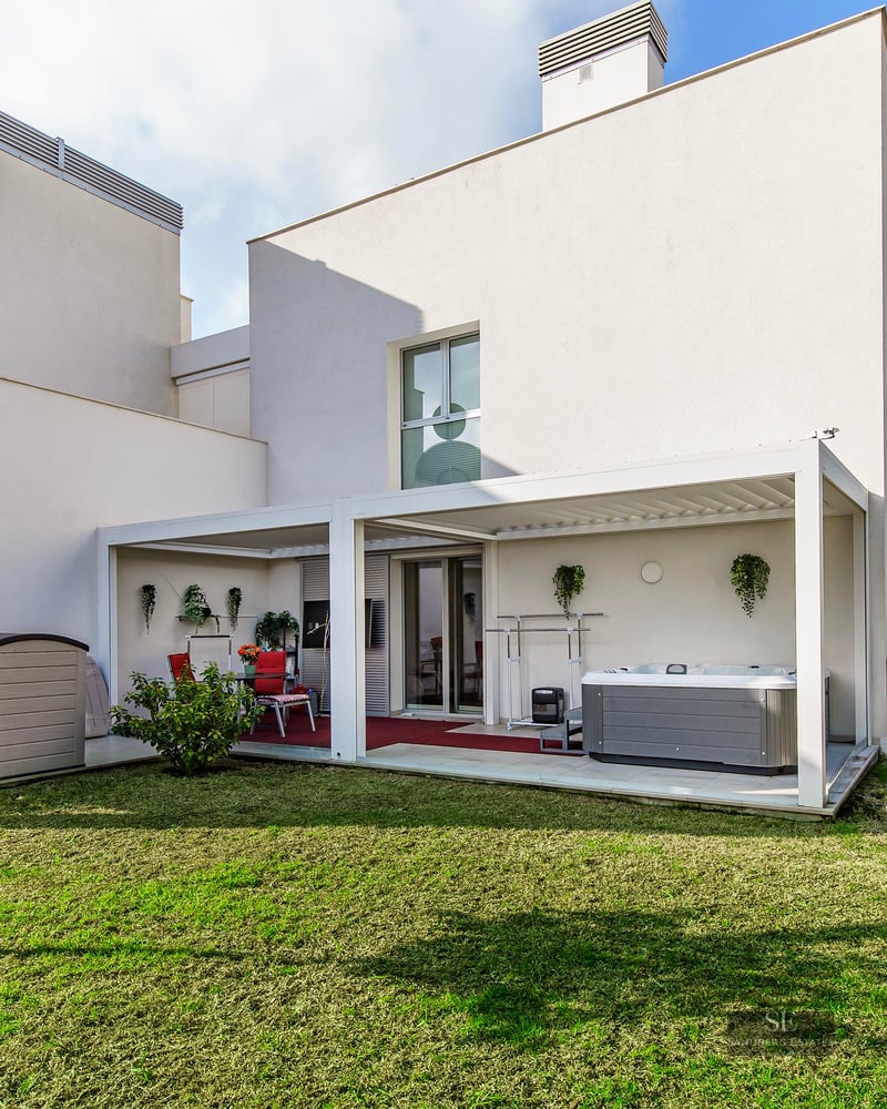 A modern white building with a green lawn and a covered terrace area featuring a hot tub and dining set.