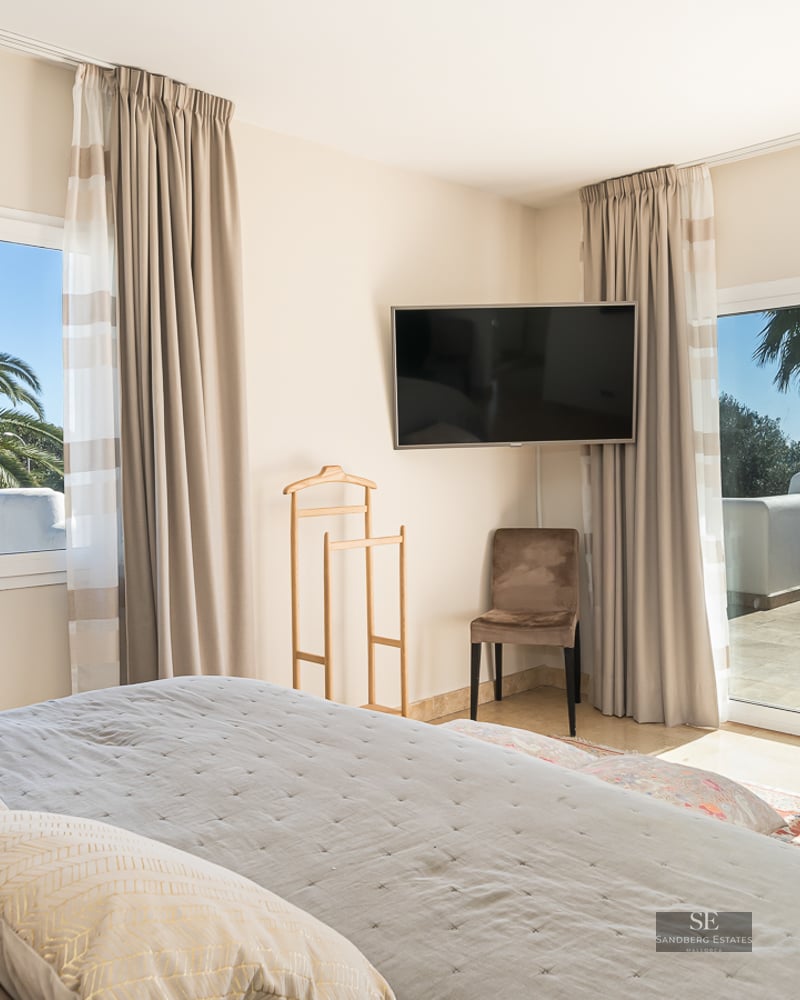 Modern master bedroom with large bed, wall-mounted TV, and sliding glass doors opening to a sunlit terrace with sea views.