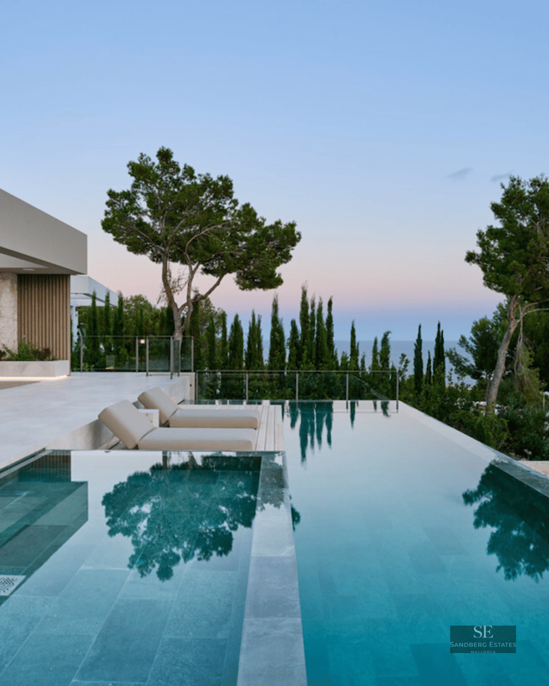 Modern infinity pool and stone terrace with lounge furniture overlooking the ocean at twilight.