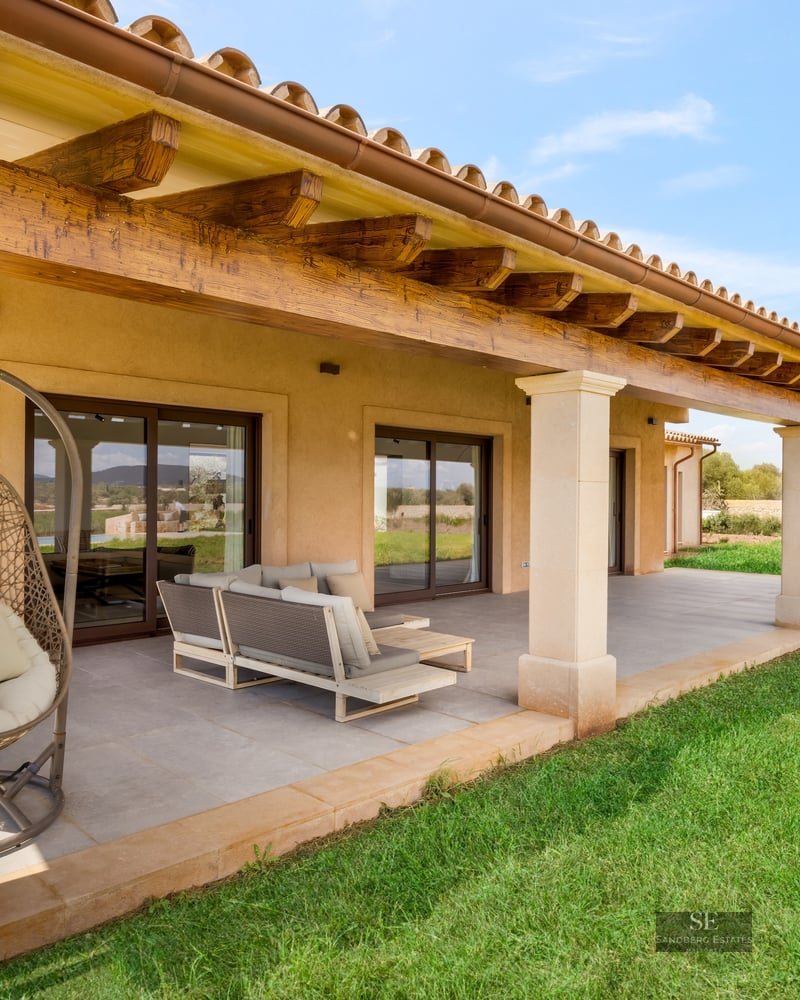 A covered terrace with rustic wooden beams, modern outdoor furniture, a hanging chair, and a green lawn under a blue sky.