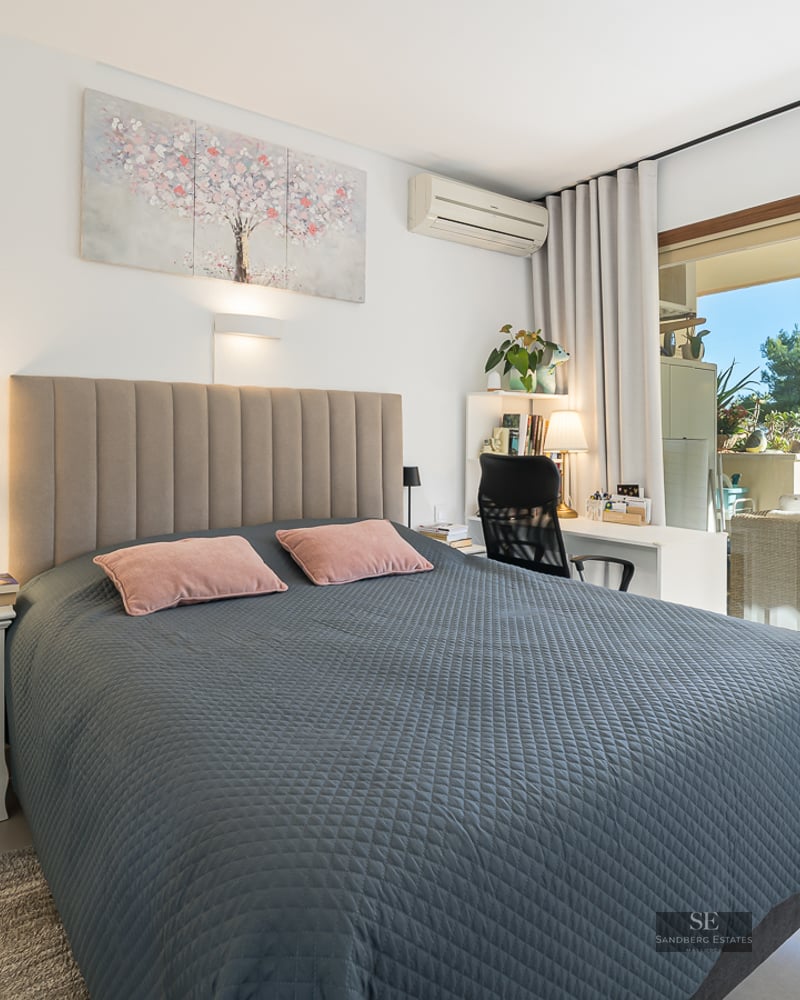 Chambre lumineuse avec lit matelassé gris, bureau et baies vitrées coulissantes ouvrant sur un balcon ensoleillé.