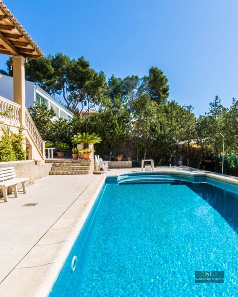 A large rectangular blue swimming pool next to a Mediterranean-style villa with a stone terrace and lush greenery.