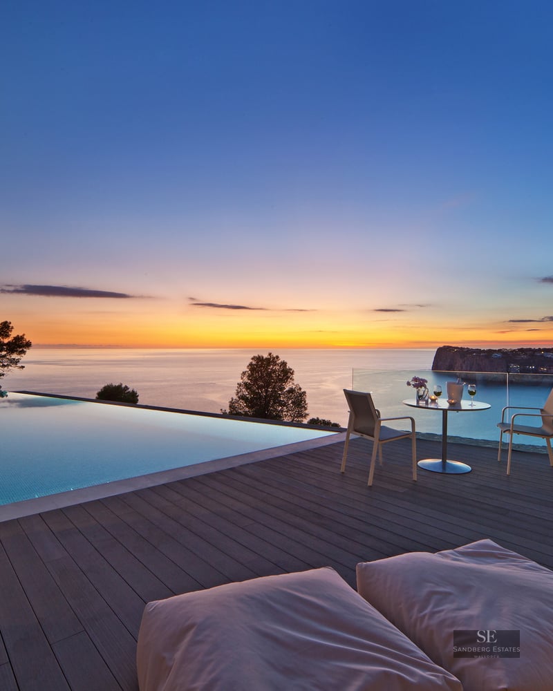 Infinity pool on a wooden deck overlooking the sea at sunset with outdoor furniture and glass railings.
