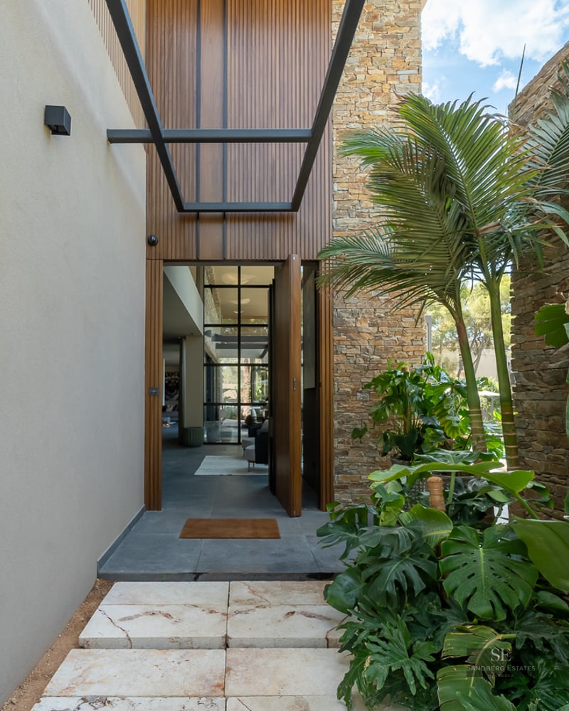 Stone path leading to a modern villa entrance with tropical plants, stone walls, and wooden door.