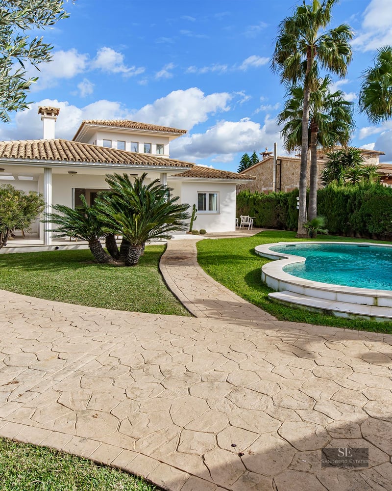A Mediterranean villa with a curved private pool, lush green lawn, palm trees, and a stone pathway under a blue sky.