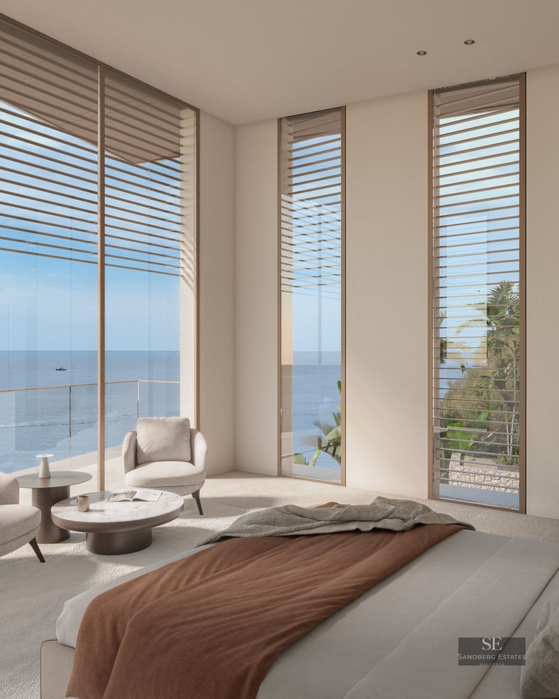 Modern bedroom with floor-to-ceiling windows overlooking the ocean and cliffs, beige armchairs, and minimalist decor.