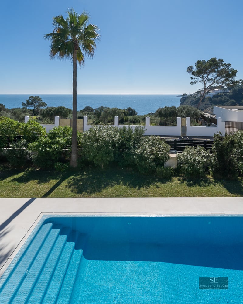 A blue tiled swimming pool and garden area overlooking the Mediterranean Sea under a clear blue sky.