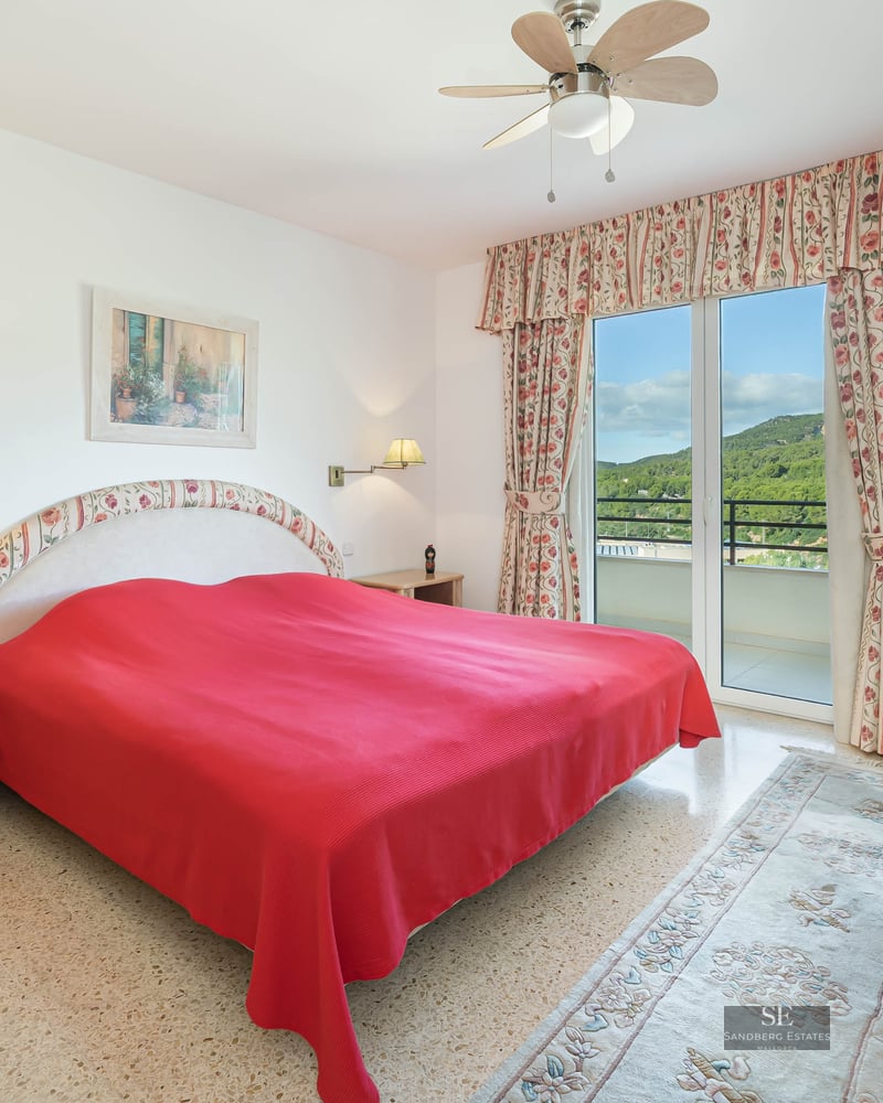 Bright bedroom featuring a red bedspread, floral curtains, and glass doors opening to a balcony with green hill views.
