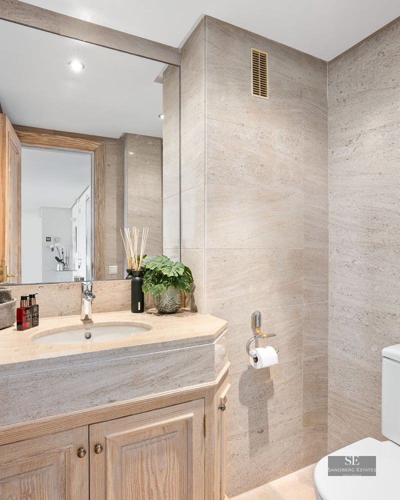 Salle de bains moderne avec carrelage en pierre beige, grand miroir et meuble sous vasque en bois.