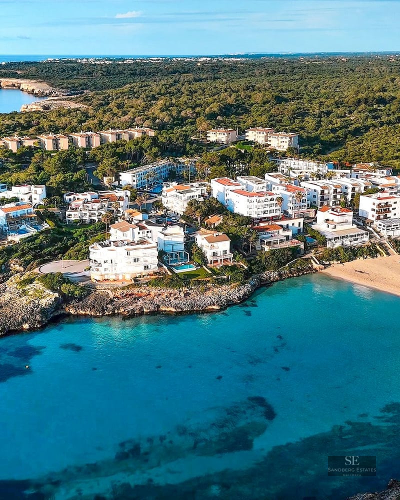Aerial view of a turquoise bay with a sandy beach and white buildings nestled between rocky cliffs and green forest.
