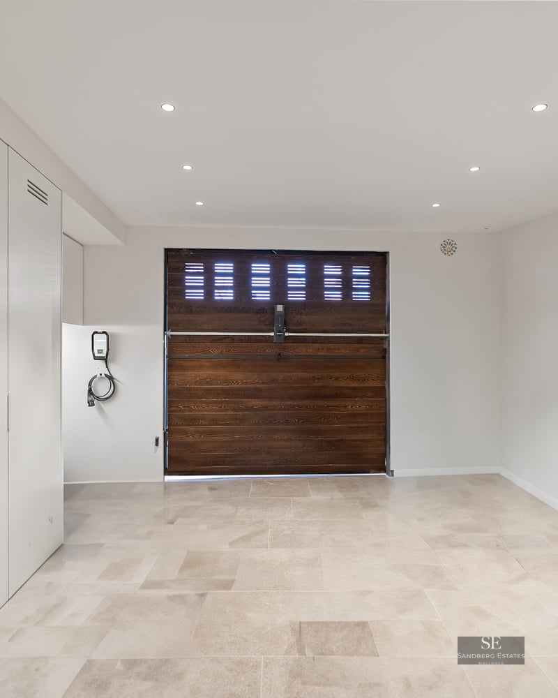 Clean modern garage with beige stone tiles, a dark wood garage door, white cabinets, and an EV charging station.